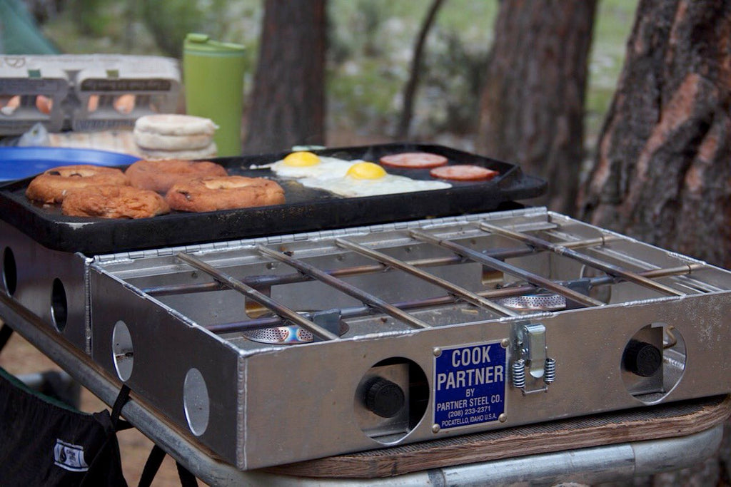 Steel 4 burner stove with a griddle on top. Cooking eggs, ham and bagel on griddle.  Background with trees.